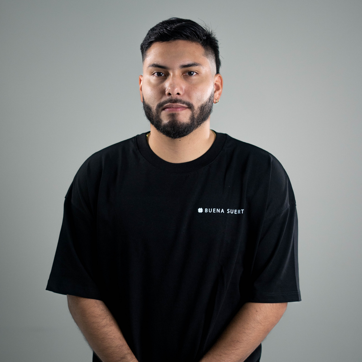 Persona de pie con camiseta negra holgada y logo blanco Buena Suerte al pecho, fondo gris
